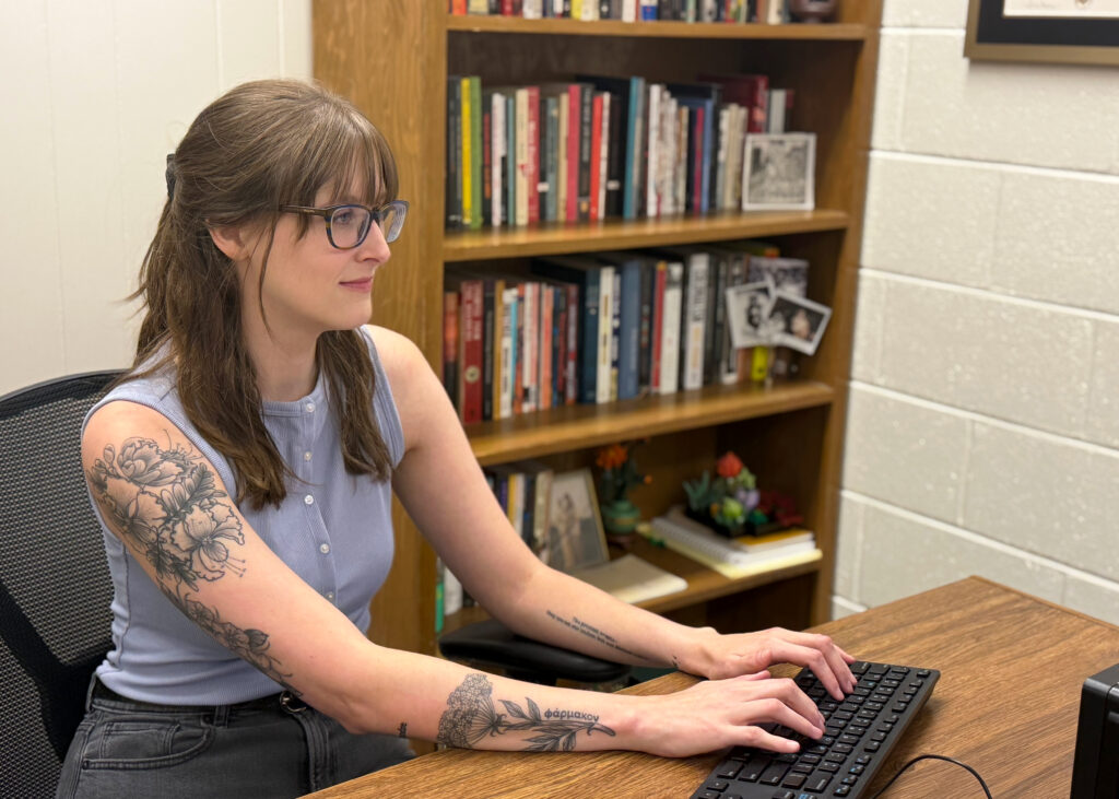 Philosophy teacher sitting at her desk typing on computer keyboard while she looks at the computer monitor. There is a book shelf full of books in the background.